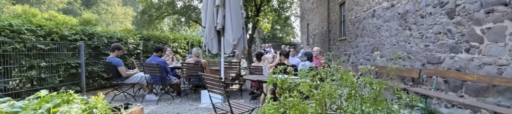 Gäste sitzen bei sonnigem Wetter an Gartentischen im begrünten Biergarten des Café Kranich am Jagdschloss Kranichstein in Darmstadt.